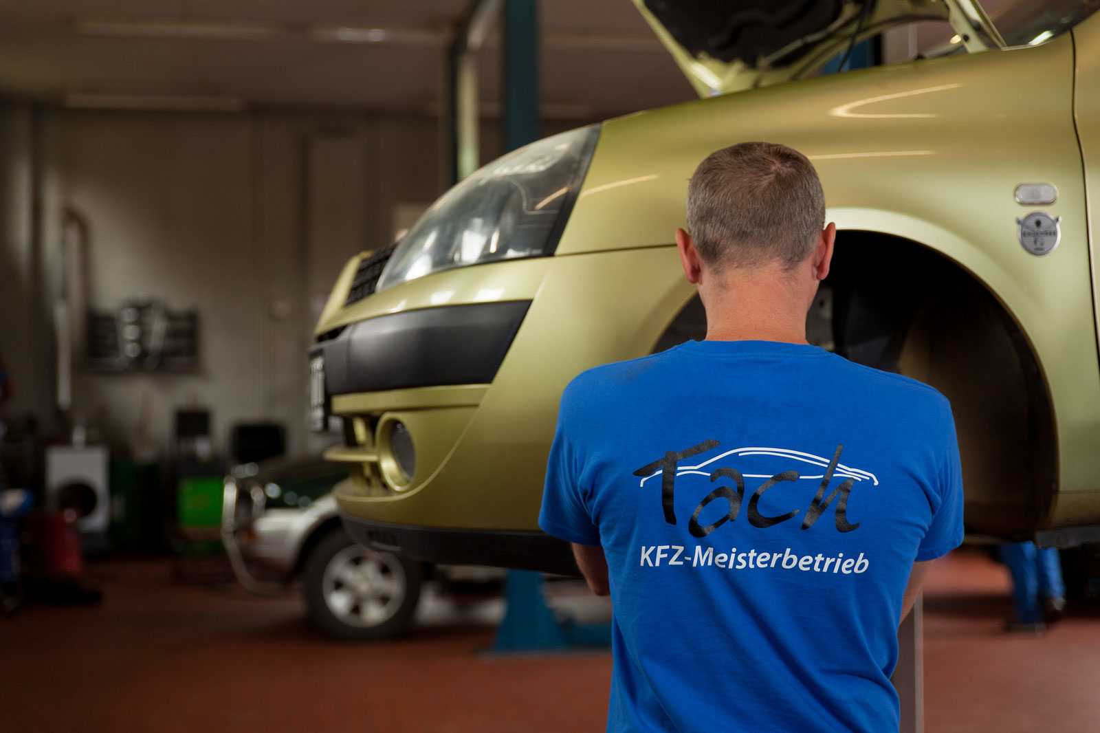 Mechaniker im Tach-Shirt vor einem Fahrzeug auf der Hebebühne